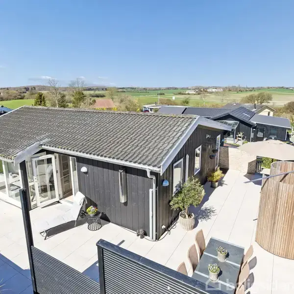 Hus med sort træfacade står på stor terrasse, omgivet af landligt landskab med marker i baggrunden. Sollys oplyser området, der har havemøbler og en rund træstruktur.