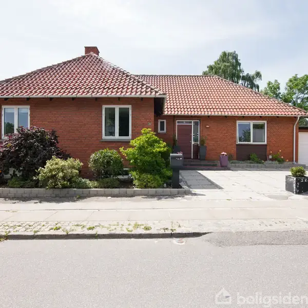 Rødt murstenshus med rødt tegltag, omkranset af velholdte buske og planter. Foran er en flisebelagt indkørsel med dekorative blomsterkasser. Solbeskinnet stille villavej i forstadsområde.