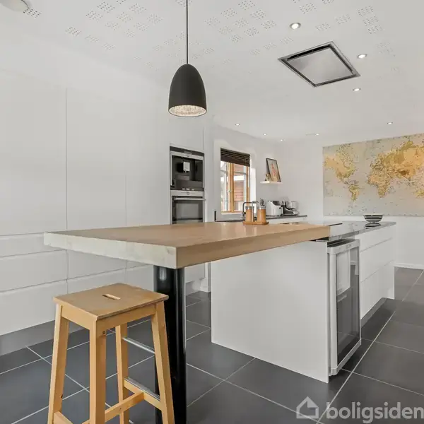 Et moderne køkken med en køkkenø og barstol, integrerede apparater og sort pendellampe. Verdenskort på væggen, flisegulv og hvide skabe skaber et minimalistisk look.