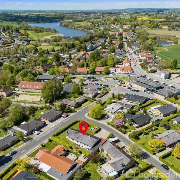 En bolig set ovenfra med et rødt markeringspunkt for placering. Huse omgiver det i en grøn forstadsområde nær en sø med åbne marker i baggrunden.