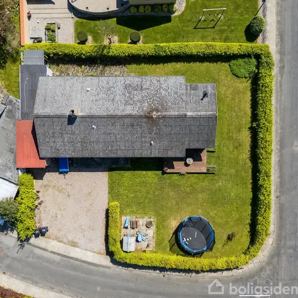 En villa med gråt tag omgivet af en hæk, beliggende i en forstad. Foran huset ses en trampolin på en velplejet græsplæne. Veje omringer ejendommen på to sider.