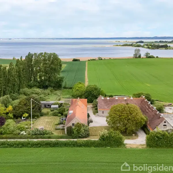 En gård ligger omgivet af grønne marker og træer, tæt på en stor sø i baggrunden. Veje og enge omgiver området i det landlige landskab.