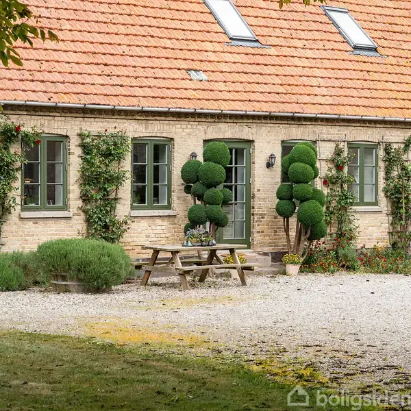 Hus med rødt tegltag og klatreplanter langs murene. Foran står formklippede buske og et træbord- og bænkesæt på grusbelagt område. Græsplæne og træer omgiver stedet.