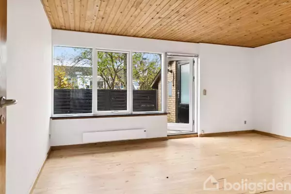 Stue med trægulv og træloft, står tom. Store vinduer og en dør fører ud til en terrasse. Lyse vægge giver atmosfære af rummelighed.