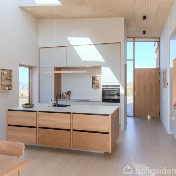 Et moderne køkken med en central køkkenø i træ. Sollys strømmer ind gennem ovenlysvinduer, der lyser de skarpe, hvide vægge op. Entrédøren ses i baggrunden.