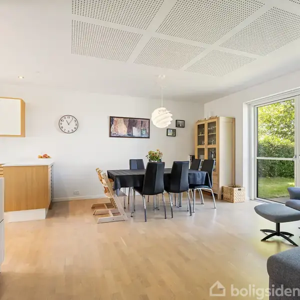 Stue med spisebord og sorte stole, sofa og lænestol til højre. Store vinduer viser grøn have. Trægulv, hvide vægge, indbygget reol, ur og kunstværker på væggen.
