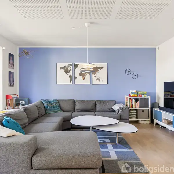 Stor grå hjørnesofa i moderne stue med hvidt loft og trægulv. Verdenskort på blå væg og fladskærm på hvidt TV-bord. Store vinduer giver naturligt lys.