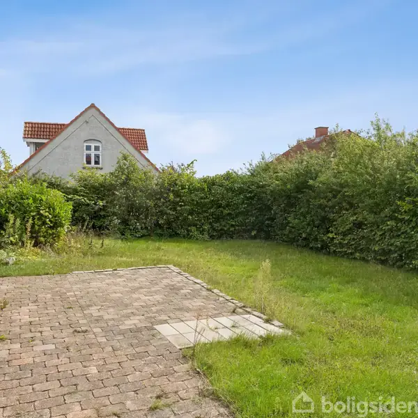 En stenbelagt terrasse ligger stille i en grøn have omgivet af tætte buske. Et hus med rødt tegltag ses i baggrunden under en lys blå himmel.