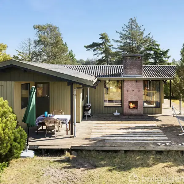 Sommerhus med træterrasse rummer spisebord og parasol, omgivet af grøn natur og træer, samt en hængekøje. En åben pejs er synlig gennem vinduet.