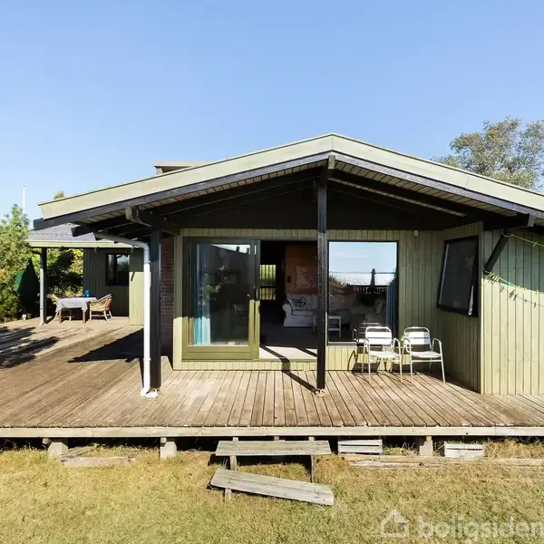 Et træsommerhus i grønlige nuancer med åbne vinduer står på en træterrasse. To stole er placeret ved glasskydedøren. Omgivet af grønne træer under en klar blå himmel.