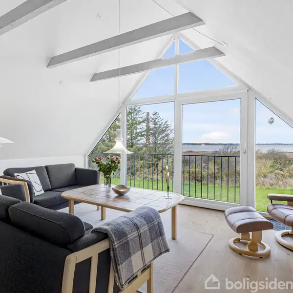 En moderne stue med sorte sofaer og en lænestol ved siden af et stort trekantet vindue. Udenfor ses en grøn have og horisonten. En loftslampe og sofabord fuldender rummet.