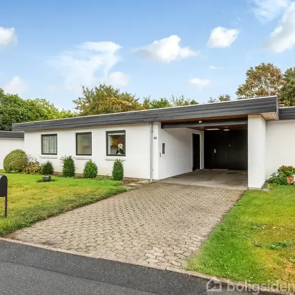 Et hvidt, moderne etplanshus står stille. Det har en indkørsel i brosten og en velplejet forhave med grønne buske. Huset ligger i et beboelsesområde med træer i baggrunden.