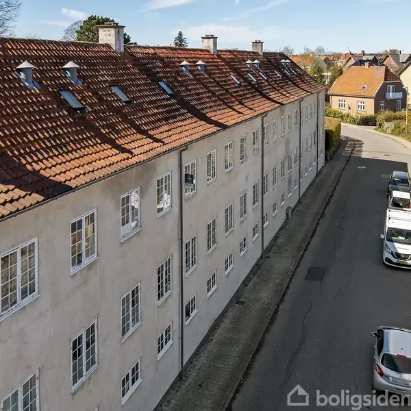 En lang bygning med rødt tegltag og hvide vinduer står langs en stille gade. Flere biler er parkeret langs vejen, og området er omgivet af andre huse.