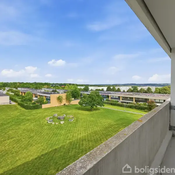 Balkon med udsigt over grøn græsplæne, hvor der står runde borde og stole. Omkring plænen ses lave bygninger og træer under en blå himmel med spredte skyer.