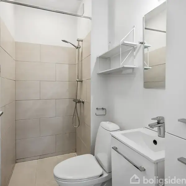 Toilet i et lille badeværelse. Håndvask med spejl og hylder ved siden af, og en bruser i hjørnet. Lyse fliser pryder væggene, skaber et moderne udtryk.