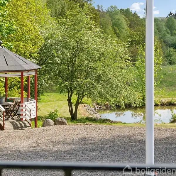 Pavillon med stole står stille i en grøn have nær en lille dam. Området er omgivet af frodig vegetation og træer.
