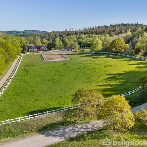 En stor, grøn indhegnet mark med en grussti langs venstre side fører til et hus i baggrunden omgivet af skovklædte bjerge.