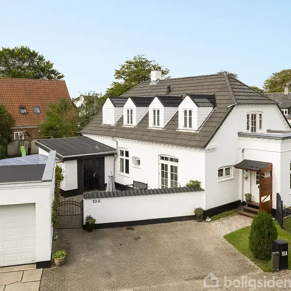 Hvidt hus med sort tag har flere vinduer, indkørsel og garage. Omgivet af have i et beboelsesområde. Tekst på husnummer er "10A" ved hoveddøren.