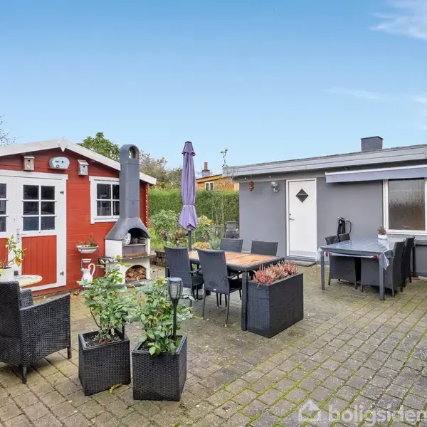 Et hyggeligt gårdhaveområde med spisebord og stole står på en flisebelagt terrasse. En rød tilbygning med skorstenspejs og en grå bygning omgiver området, der har blå himmel.