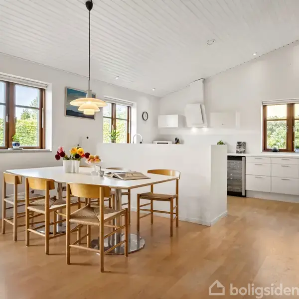 Et moderne køkkenalrum med et hvidt spisebord og seks træstole, blomsterdekoration, og en moderne loftslampe. Rummet er lyst med hvide vægge og store vinduer, der lukker naturligt lys ind.