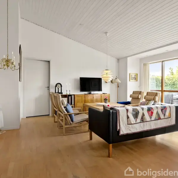 Stue med sort sofa centralt placeret, hængende lys over bordet, TV på reol, store vinduer med udsigt til terrasse og have. Trægulv og lyse vægge skaber en moderne atmosfære.