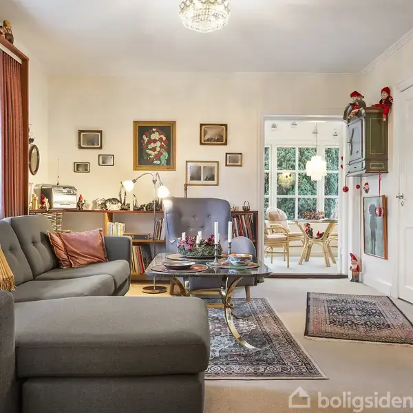 Grå sofa med puder står stille i et hyggeligt stueområde, dekoreret med billeder på væggen og julepynt. I baggrunden ses ind til en spisestue med vinduer og planter.