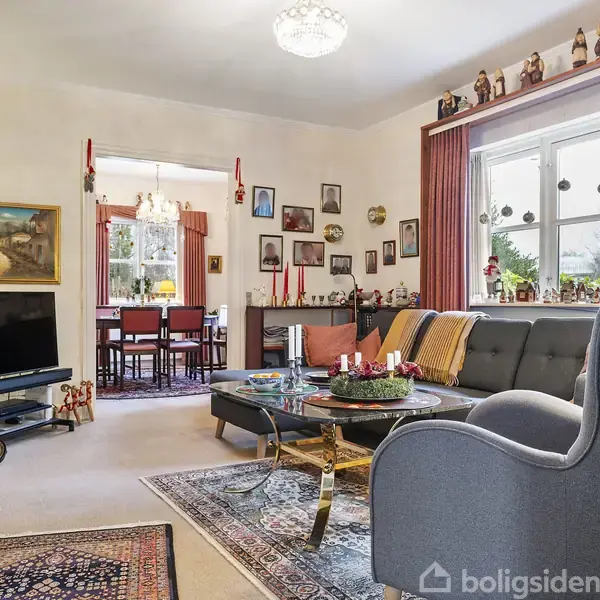 En stue med sofamøbler og sofabord, dekoreret med røde gardiner og billeder på væggene. I baggrunden ses en spisestue med et bord og stole.