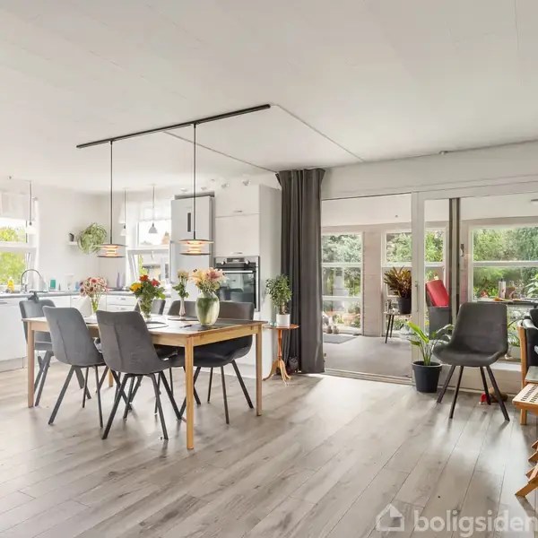 Spisebord med stole står i midten af et lyst køkken-alrum. Køkkenet har hvide skabe og vinduer til venstre. Store glasdøre fører til terrassen og haven udenfor.
