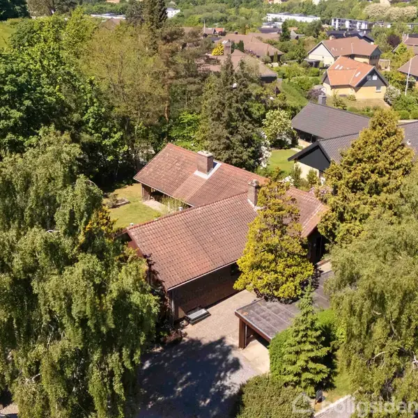 Hus med rødt tegltag placeret blandt frodig, grøn vegetation. Omkring huset er en villakvarter med flere huse. Solskin spreder lys over området, hvilket giver et sommerligt indtryk.