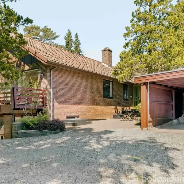En murstensvilla med tegltag er omgivet af træer og buske. Den har en tilhørende carport og en hævet terrasse med træværn. Grusbelagt indkørsel leder til huset.