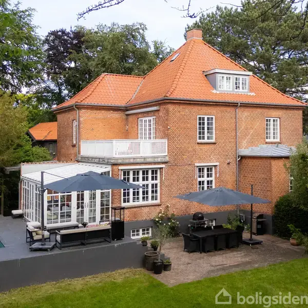 Et to-etagers hus i mursten står med orangerødt tegltag. Foran huset er en terrasse med pool, havemøbler samt parasoller. Omgivelserne består af grønne træer og græsplæne.