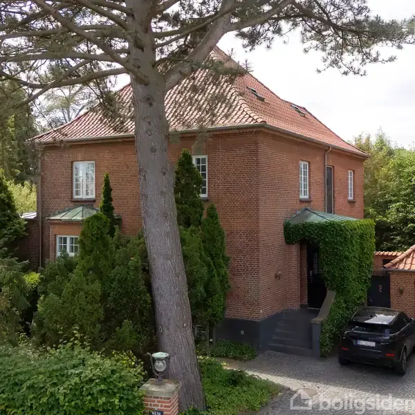 Et rødt murstenshus med skråt tag står omkranset af store træer og grøn vegetation. En sort bil holder parkeret ved husets garage, der er delvist tildækket af klatreplanter.
