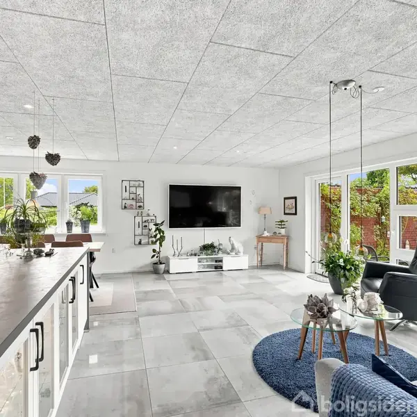 Et moderne stueområde med lyse flisegulve og store vinduer. En sort lænestol står ved et rundt blå tæppe, mens planter og dekorationer pryder rummet. En stor fladskærm hænger på væggen.