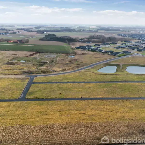 Gule marker strækker sig ud med asfalterede veje, der skærer gennem landskabet. To små søer ligger ved siden af vejene. I baggrunden ses en bebyggelse og landlige omgivelser.