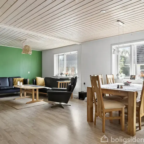 Stue med spisebord og stole til højre, og sofa-sæt med borde til venstre. Grøn væg og træ-loft skaber en hyggelig atmosfære. Store vinduer lader naturligt lys strømme ind.