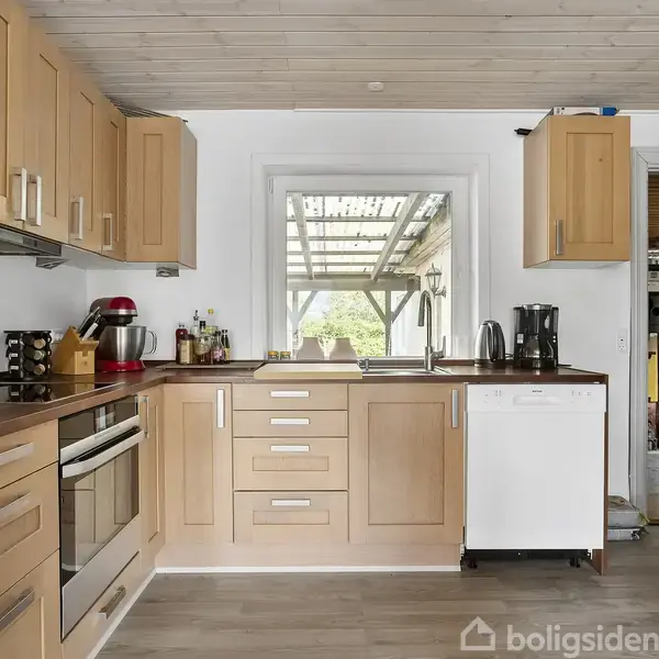 Køkken med træskabslåger og hvidt opvaskemaskine, kaffemaskine og røremaskine på bordet. Et vindue viser en overdækket terrasse udenfor. Dør til højre fører til et andet rum.