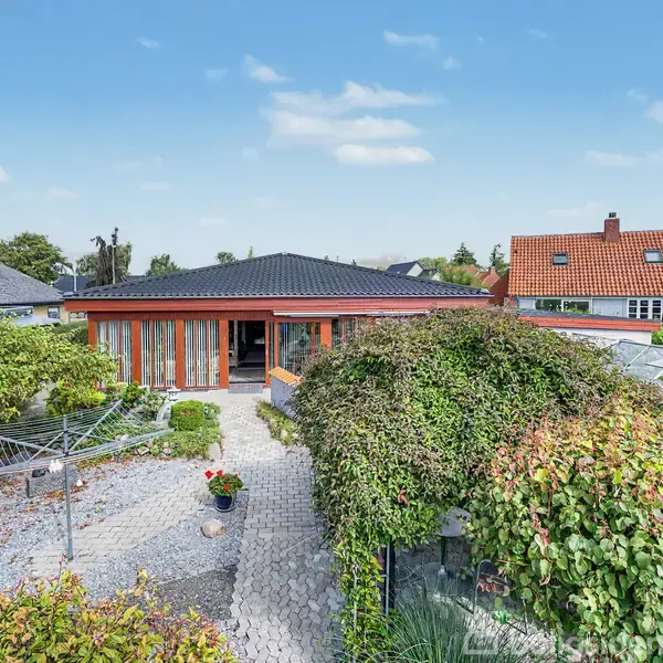 Et moderne hus med rødt tag står omgivet af en frodig have. En flisebelagt sti fører gennem haven, der har en glasdrivhus og velfriserede buske.
