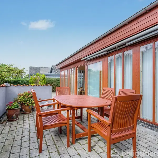 Et rundt træbord med seks tilhørende stole står på en flisebelagt terrasse. Omkring står blomsterkrukker, og en bygning med store vinduer danner baggrunden.