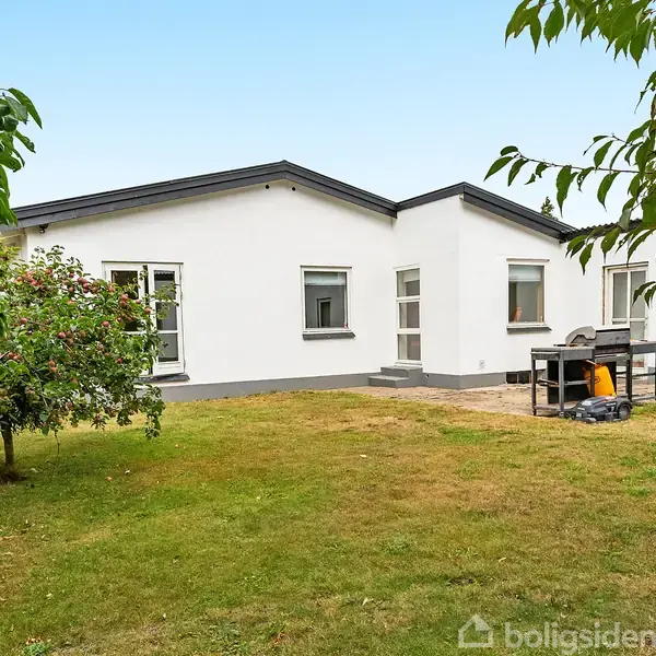 Hvidt hus står i en frodig have med græsplæne. To åbne vinduer vendt mod haven. Havegrill og en lille terrassemur er til højre.