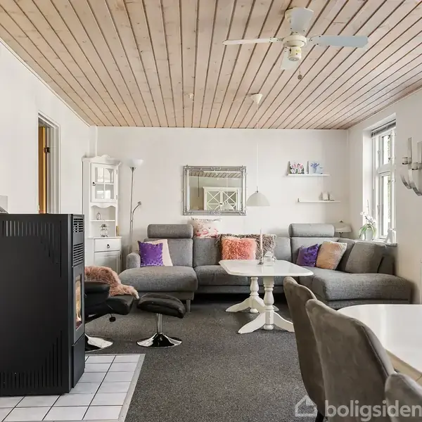 Stue med grå sofaarrangement under træloft. Pejs ved flisebelagt område. Vinduer giver naturligt lys. Et spisebord i forgrunden og diverse hylder og dekorationer pryder væggene.