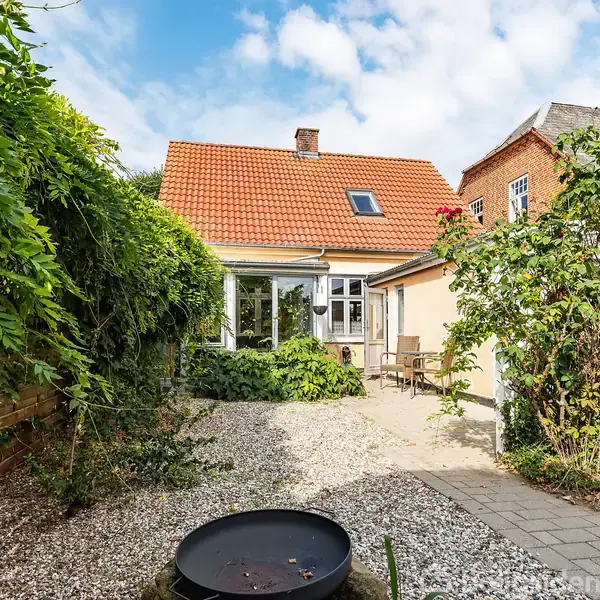 Hus med rødt tegltag og hvide vinduer står i solskinsbelyst gårdhave omgivet af grønne planter. Foran huset er der grusplads med ildsted og flisebelagt terrasse med havemøbler.
