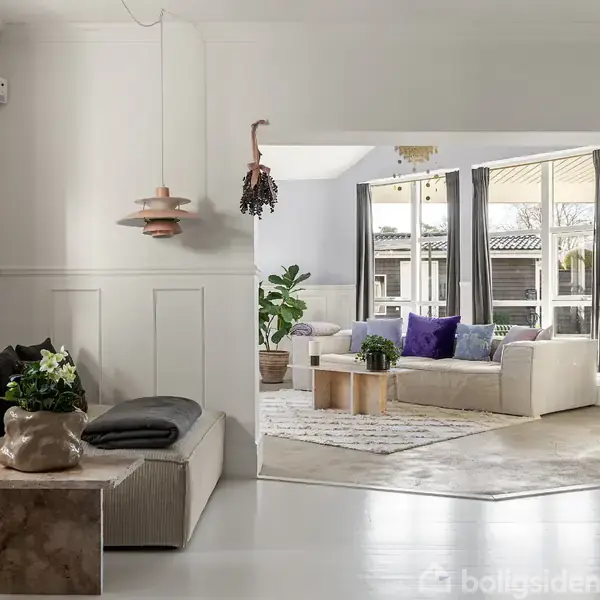 En stue med moderne indretning, hvide vægge og møbler, herunder en beige sofa med farverige puder. Store vinduer slipper naturligt lys ind, og en lille plante pynter rummet.