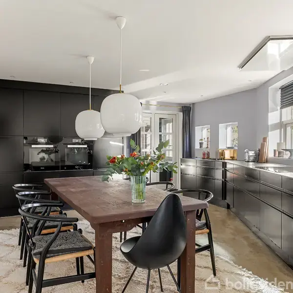 Et stort spisebord med en blomsterbuket står i et moderne køkken. Køkkenet har sorte skabe, store hængelamper og et poleret betongulv med tæppe, der skaber en elegant atmosfære.