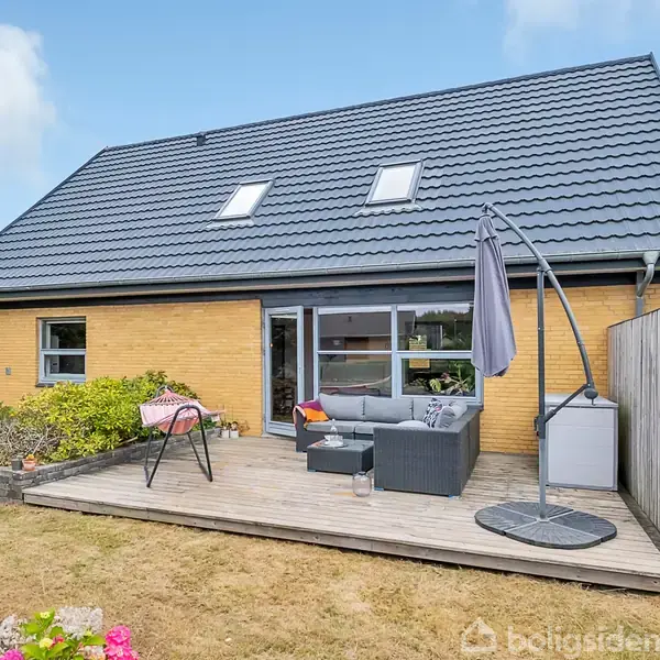 Huset er murstensbygget med et mørkt, skråt tag. En trækterrasse har loungemøbler og en paraply, der skaber en hyggelig udendørs atmosfære. Plænen omkring er grøn med blomster.