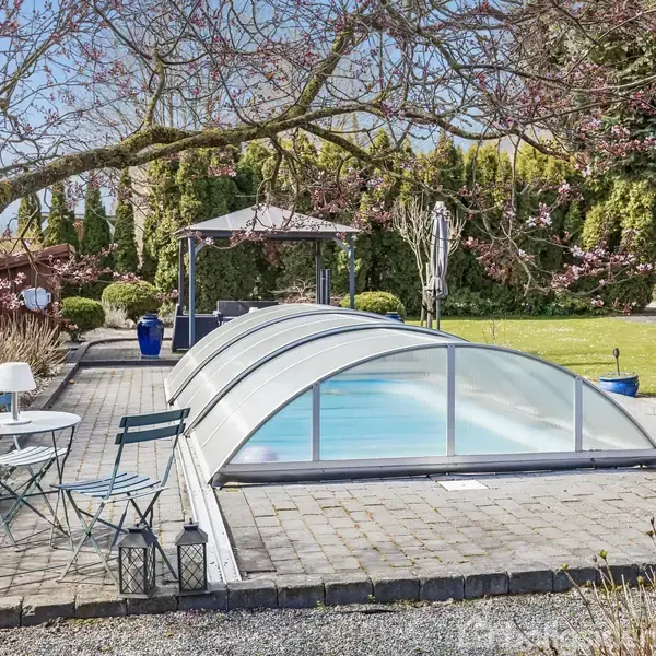 Et overdækket swimmingpool sidder midt i en flisebelagt terrasse med et bord og to stole ved siden af. I baggrunden ses en have med græs, træer og buske.