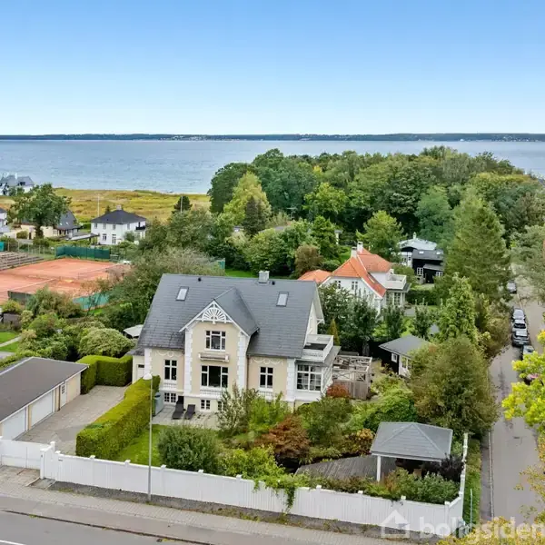 Stort hus med gråt tag omgivet af en velplejet have. Det ligger i et grønt boligområde nær tennisbaner med udsigt over en fjord eller sø i baggrunden under en klar blå himmel.