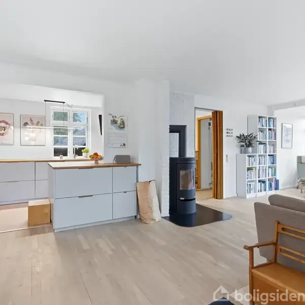 Et moderne, lyst køkkenalrum med hvide skabe, en stor køkkenø og vindue. I baggrunden ses en brændeovn, en bogreol og en lænestol ved store vinduer, der slipper lys ind.