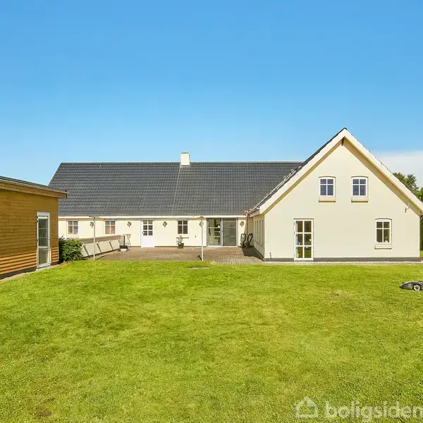 Et hvidt hus med sort tag står i en stor have med grøn græsplæne. Til venstre er et træskur. Himlen er klar og blå.