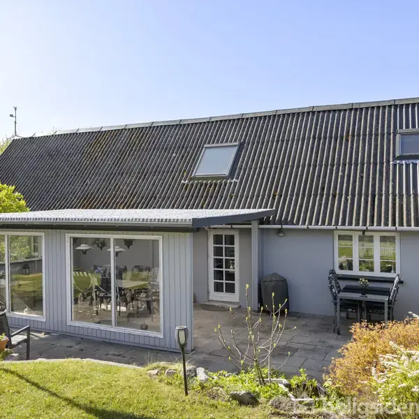 Et gråt, moderne hus med store vinduer og en dansk flagstang står i en solrig have med terrasse. Spisebord og stole er placeret på terrassen under det skråt tag.