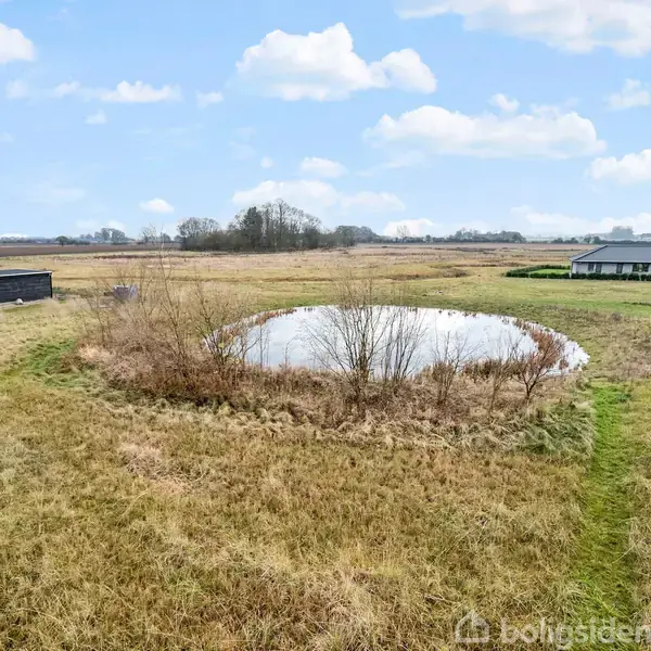 En lille sø omgivet af græs i et åbent landskab. Til venstre ses en sort bygning, og til højre nogle huse. Himlen er blå med spredte skyer.